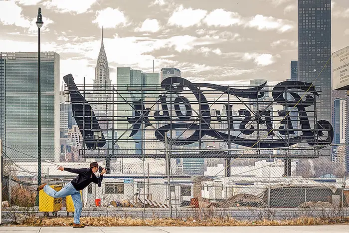 a man stands behind the Pepsi sign in Long Island City, with Manhattan in the background